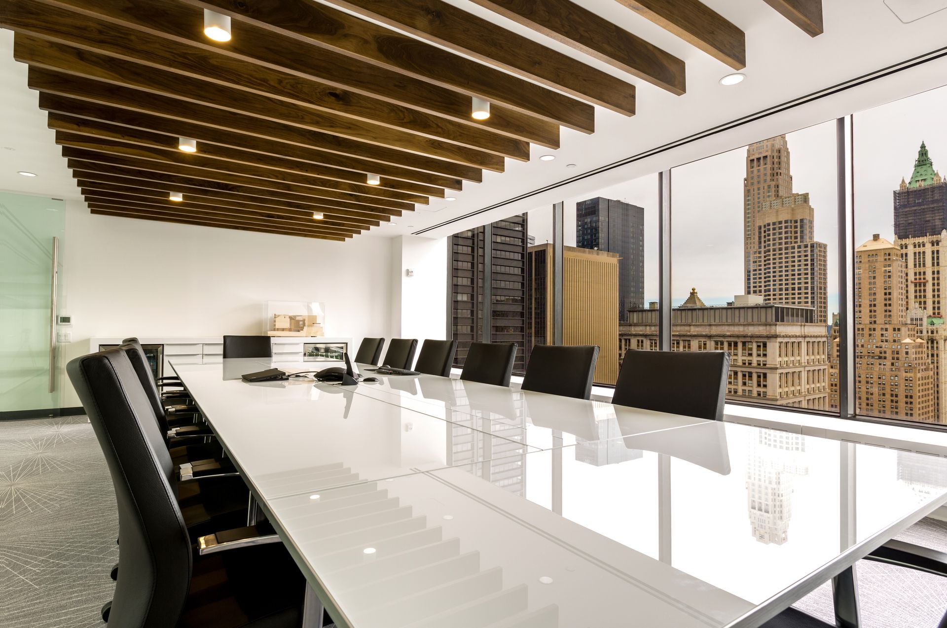 Conference room with white table and black chairs, large windows with city view, wood ceiling.