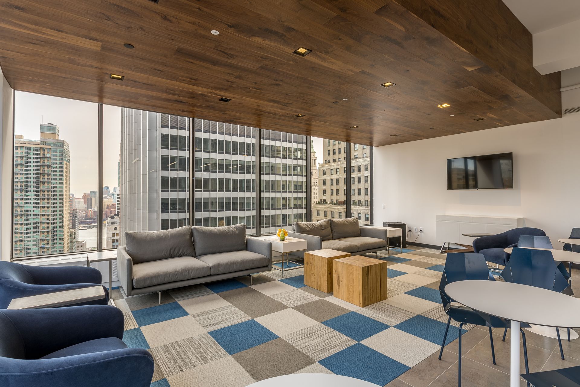 Modern lounge with city views, sofas, chairs, wooden ceiling, checkered carpet, and a mounted TV.