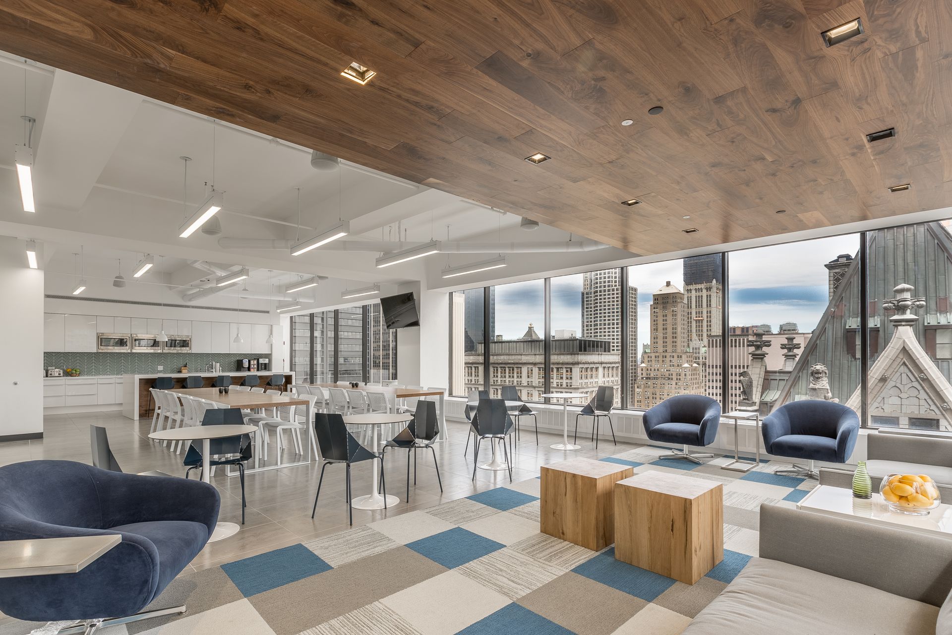 Modern office lounge area with seating, tables, kitchen, and large windows overlooking city buildings.