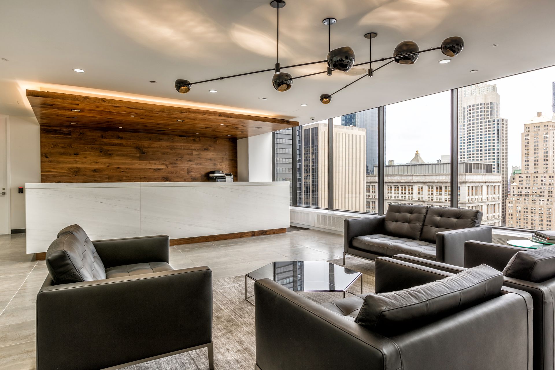 Modern office waiting area with dark gray sofas, glass coffee table, and large windows overlooking city buildings.