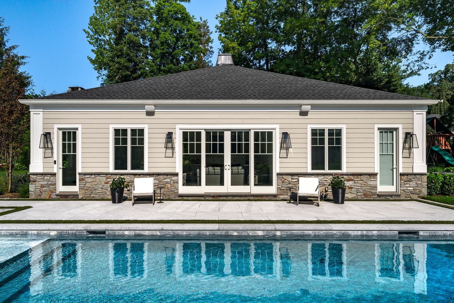 Pool house with pool in foreground; beige siding, stone base, black roof, glass doors, and windows.