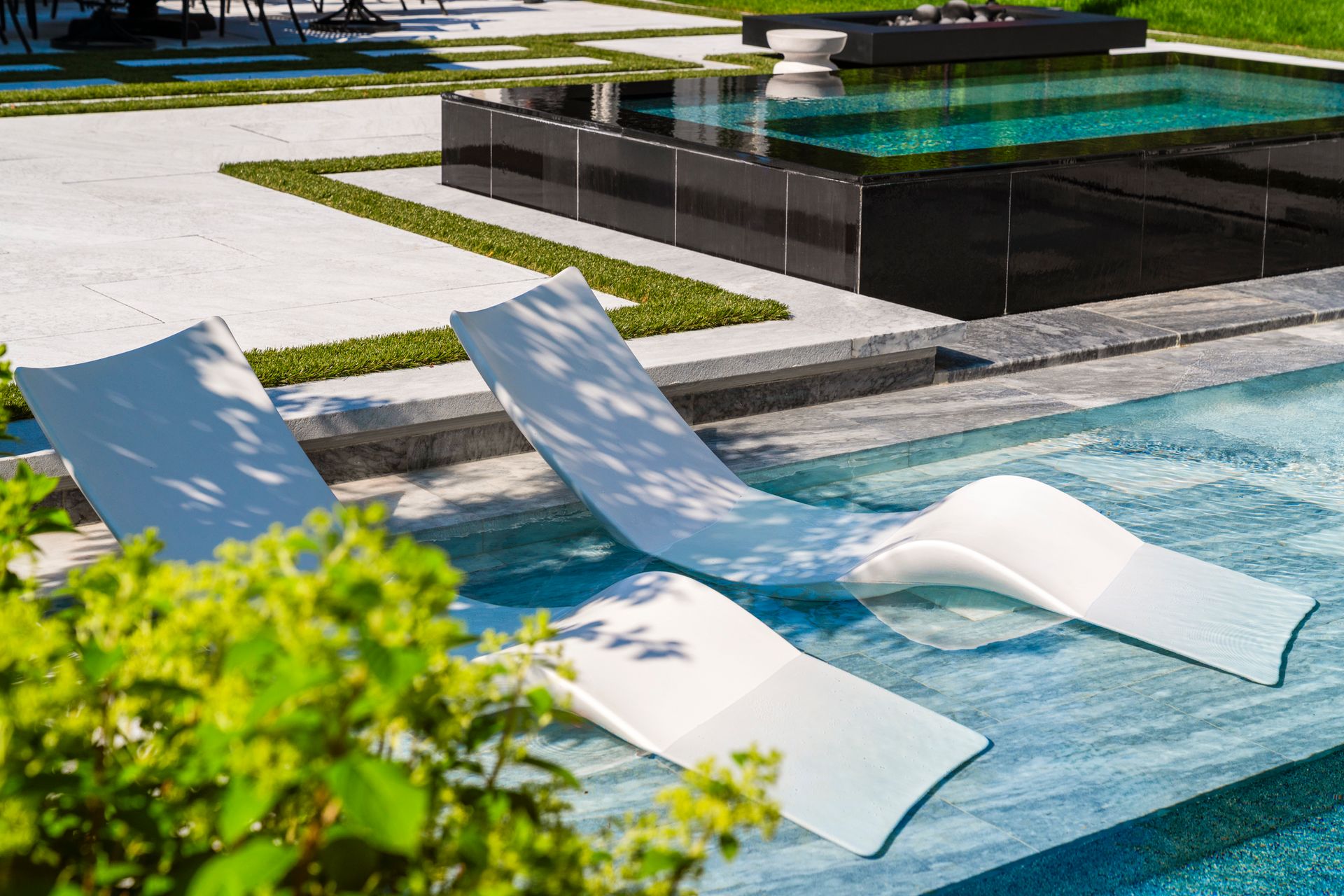Three white chaise lounges in a pool, greenery in foreground, reflecting pool in background.