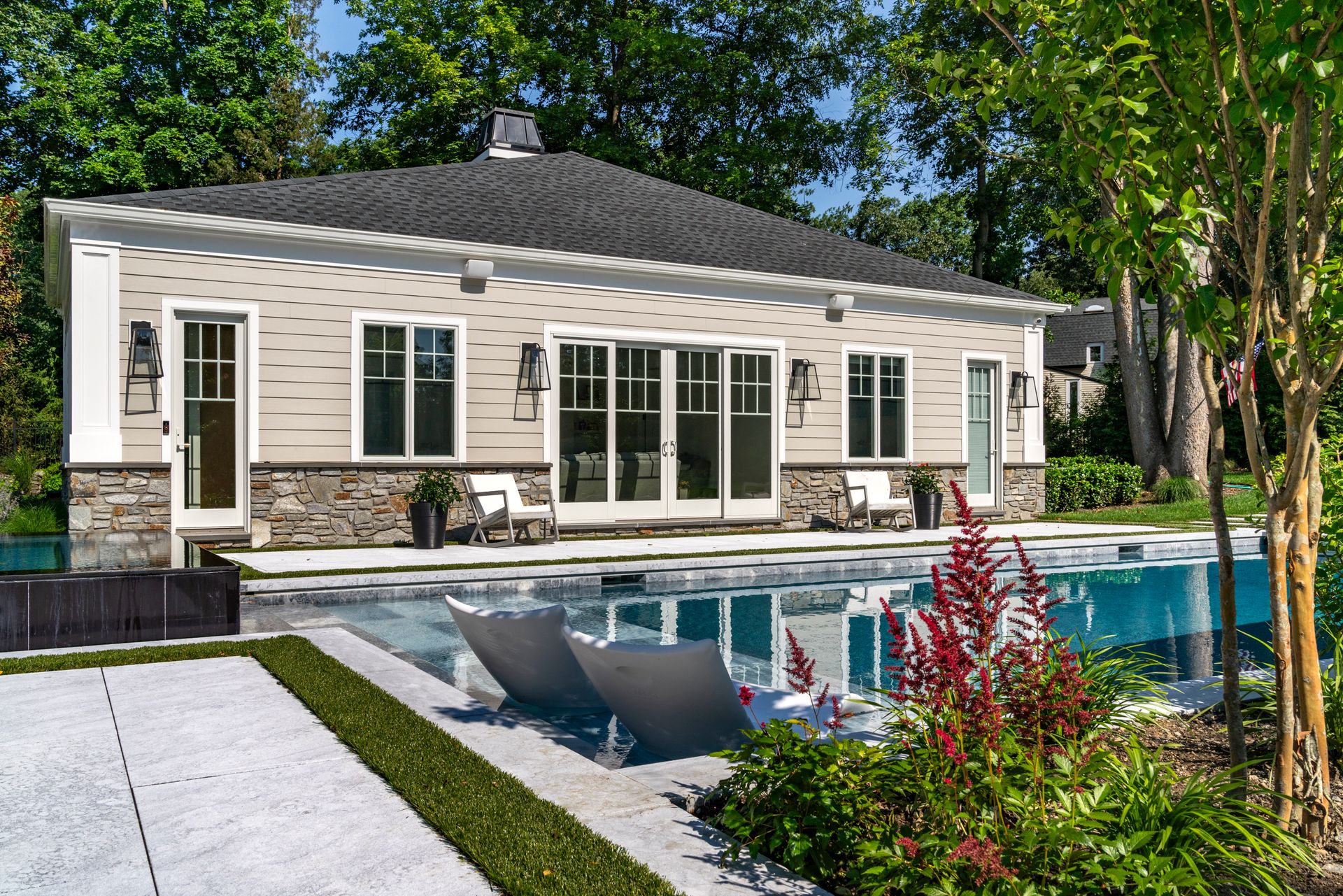 Poolside building with light-colored siding, dark roof, windows, and stone base. Reflective pool with modern lounge chairs.