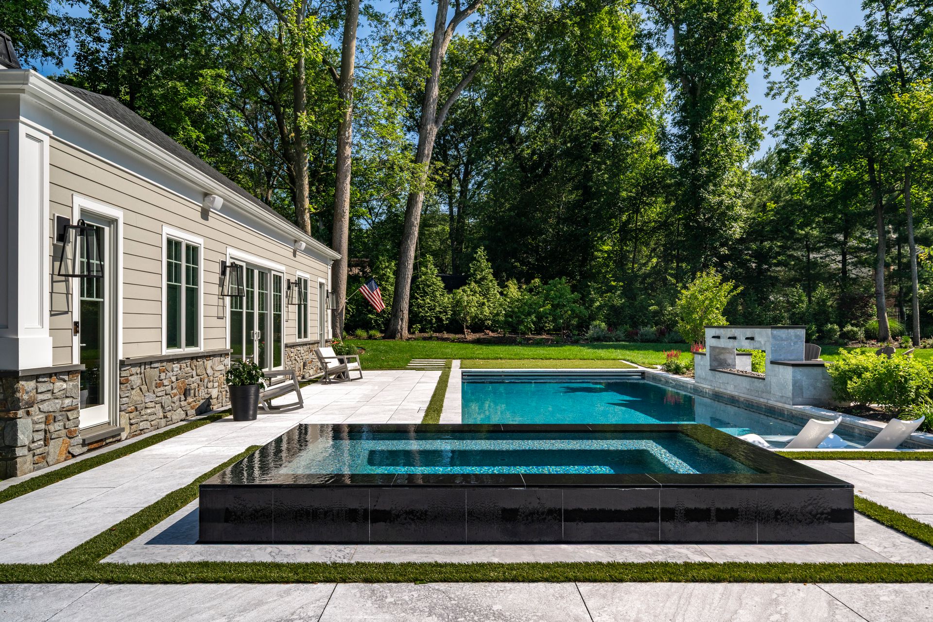 Backyard pool and spa with manicured lawn, house with stone accents, trees.