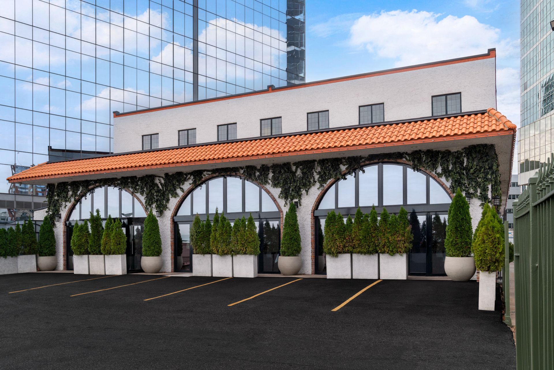 Restaurant with arched windows, terra cotta roof, and greenery, set against a glass skyscraper.