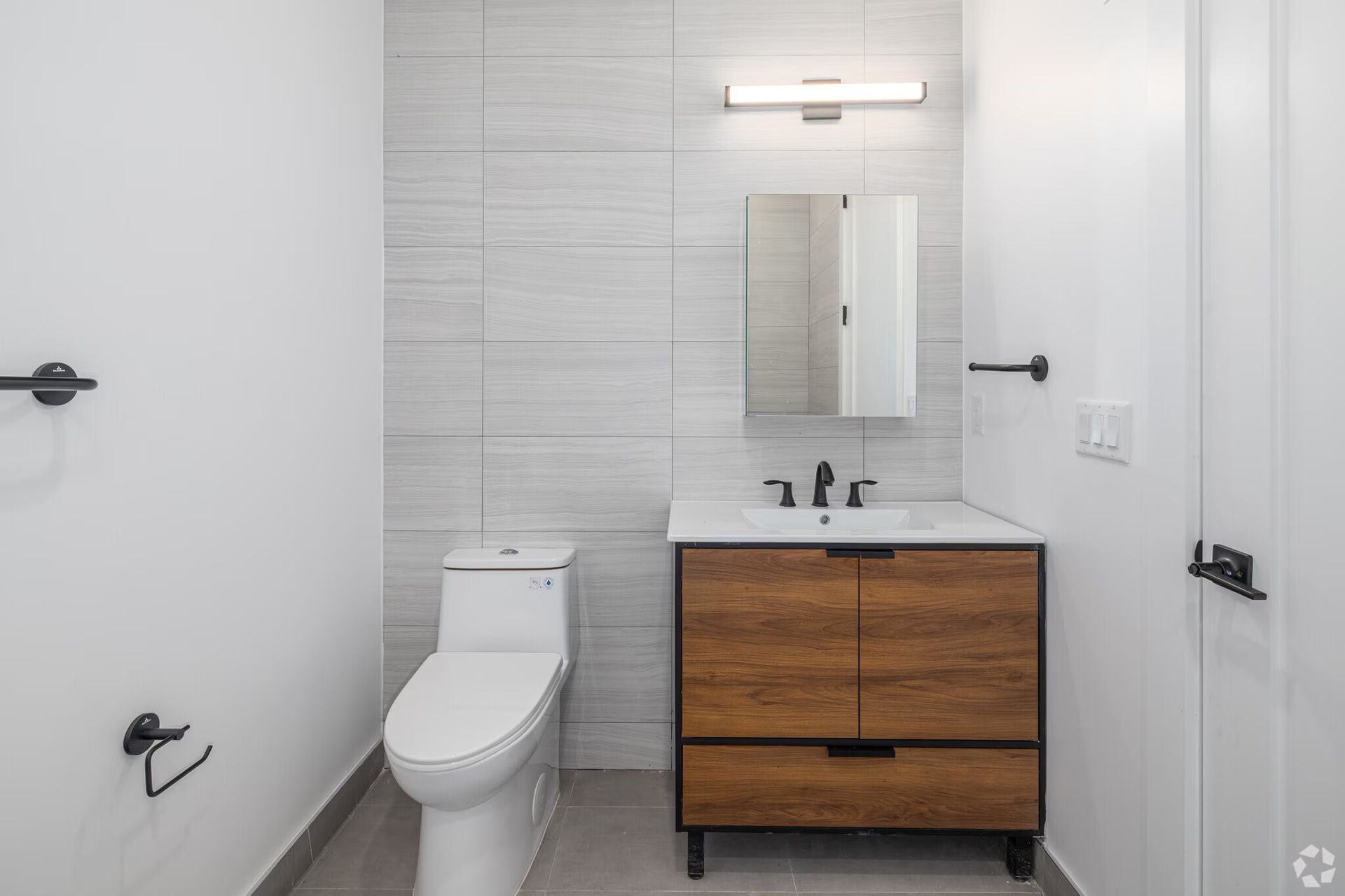 Modern bathroom with white toilet, wood vanity, and gray tile wall.