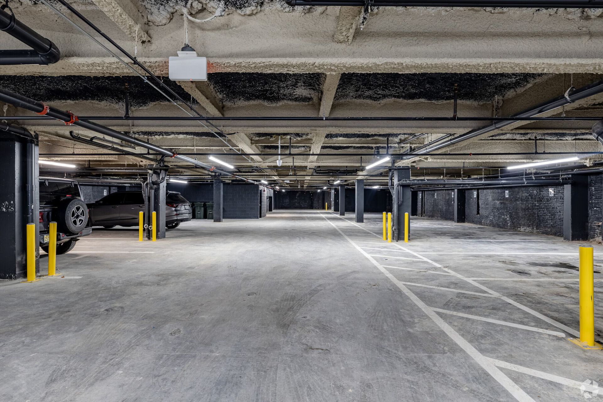 Empty parking garage with gray concrete floor, columns, and ceiling; a few cars parked near the left.