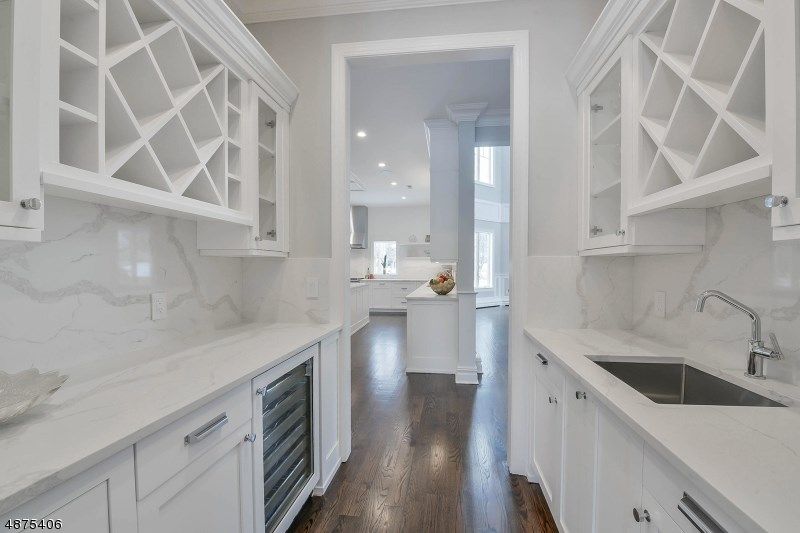 Bright, narrow kitchen space with white cabinets, wine rack, countertops, and a sink. Dark wooden floor leads to another kitchen area.