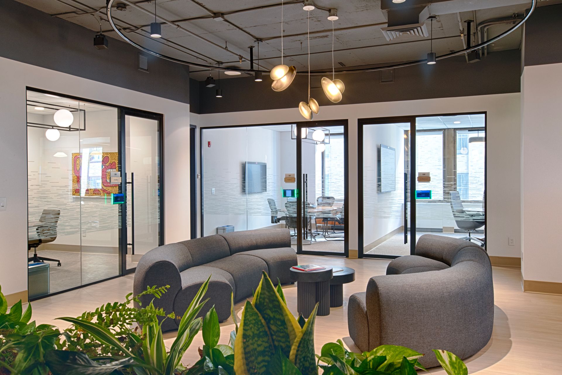 Modern office waiting area with curved gray sofa, glass-walled conference rooms, and decorative lighting.