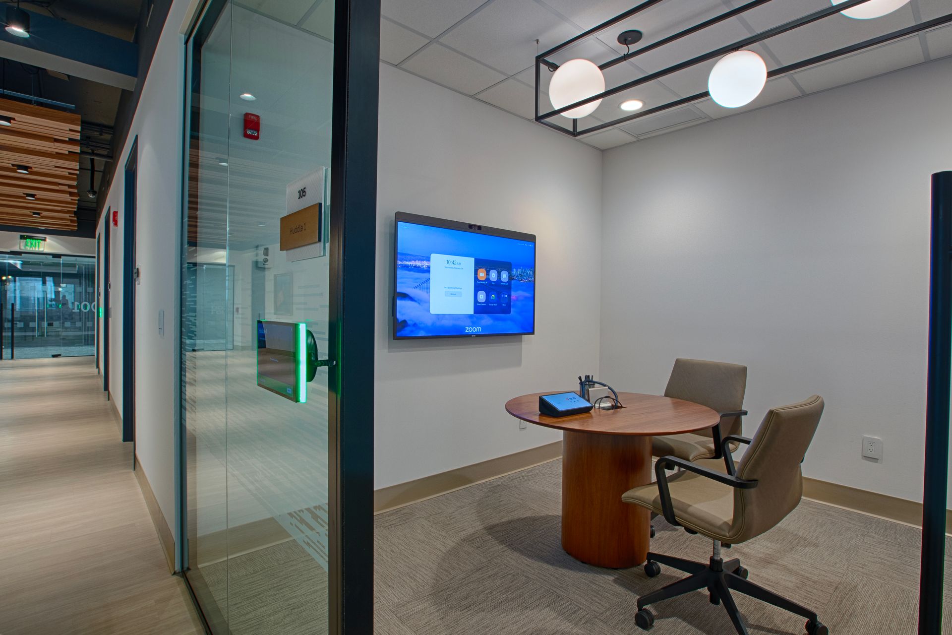 Meeting room with table, chairs, monitor, and light fixture; hallway with glass doors visible.