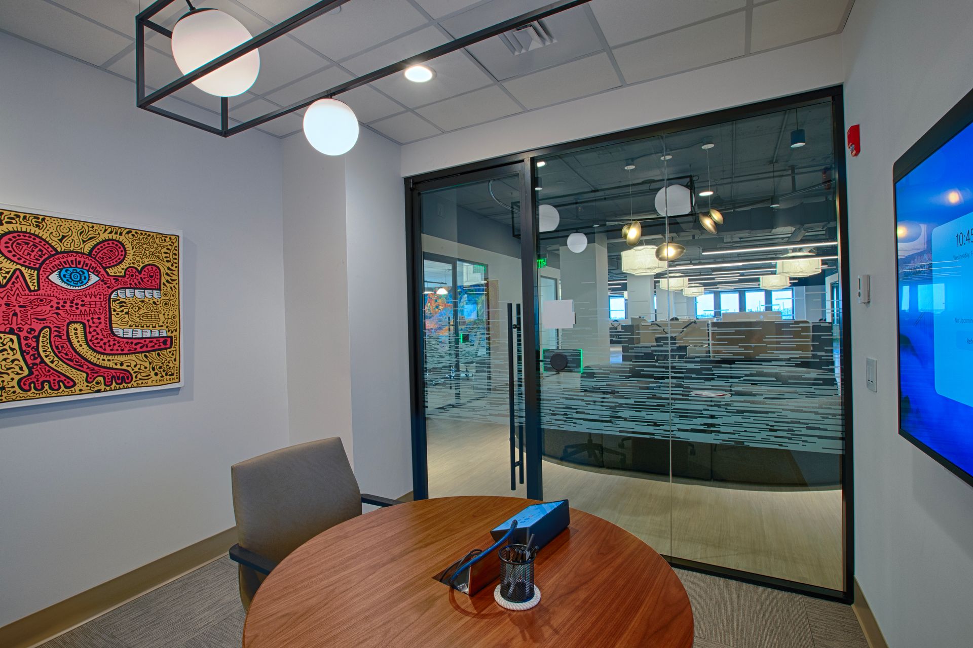 A small office with round table, artwork, and glass door, looking into a larger office space.