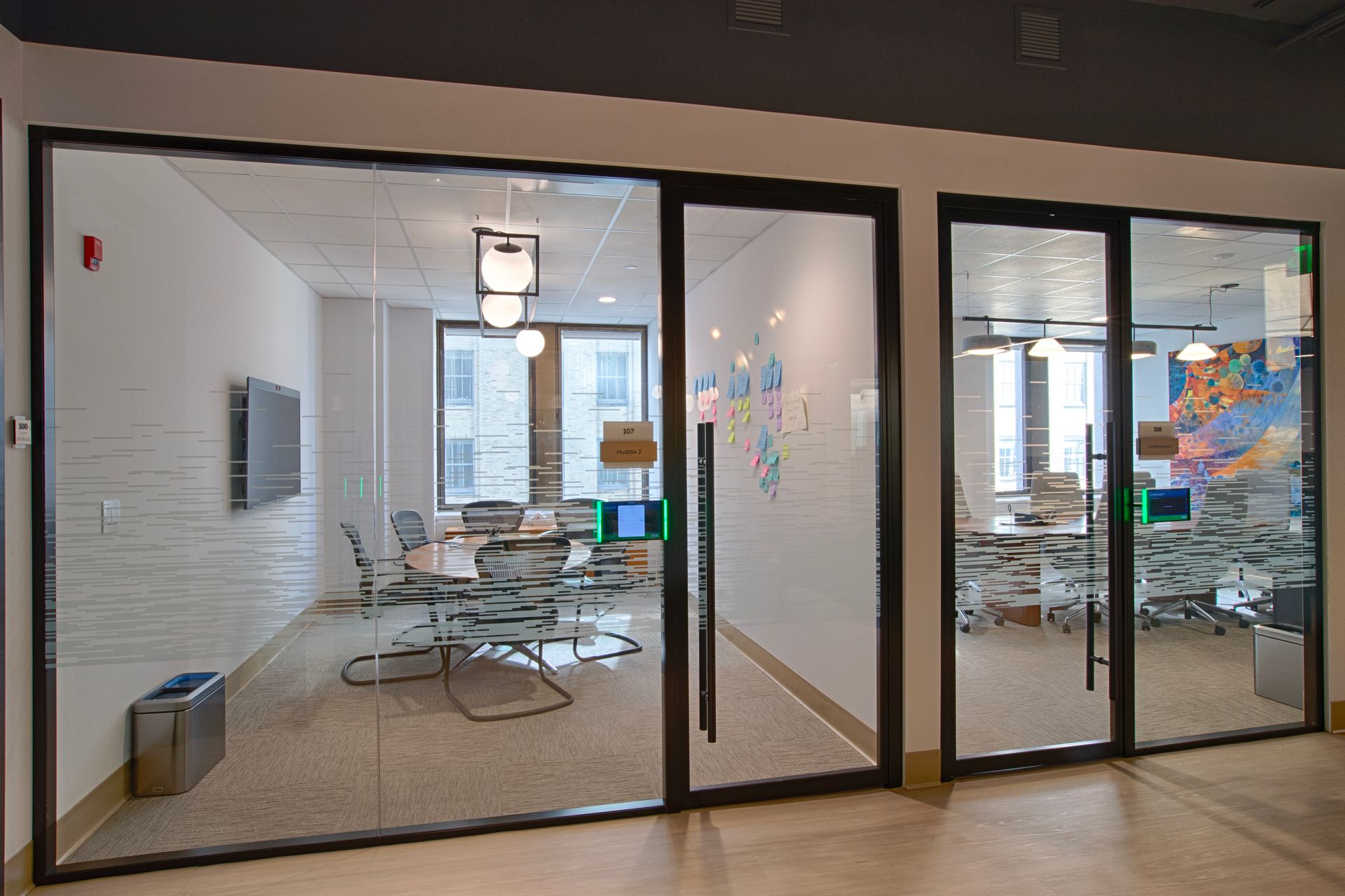Glass-walled office spaces with black frames. One room has a conference table, another has desks. Beige carpet and a gray ceiling.