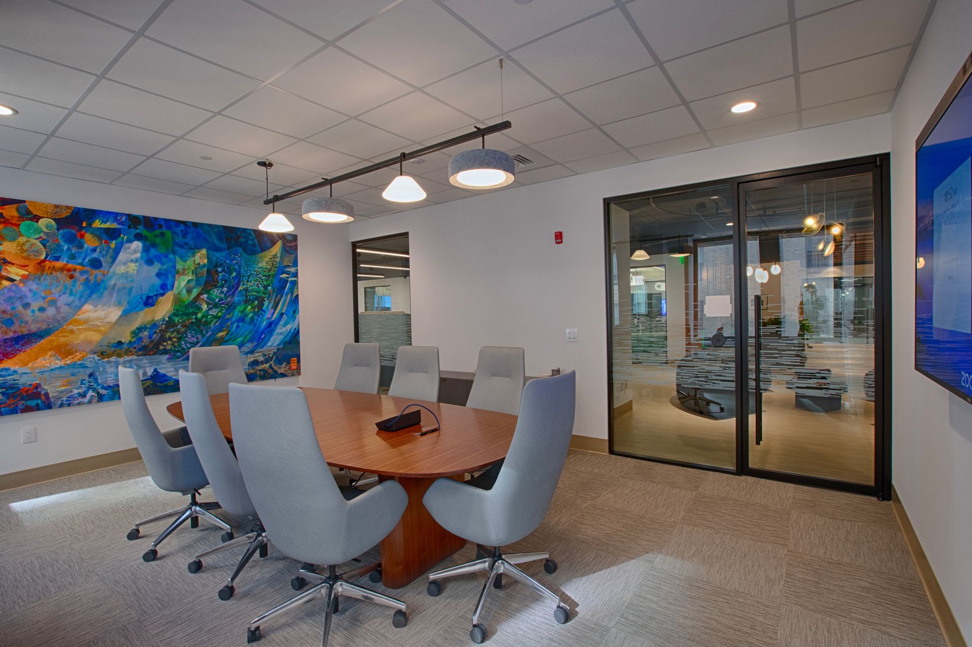Conference room with round table, gray chairs, colorful artwork, glass wall, recessed lighting.
