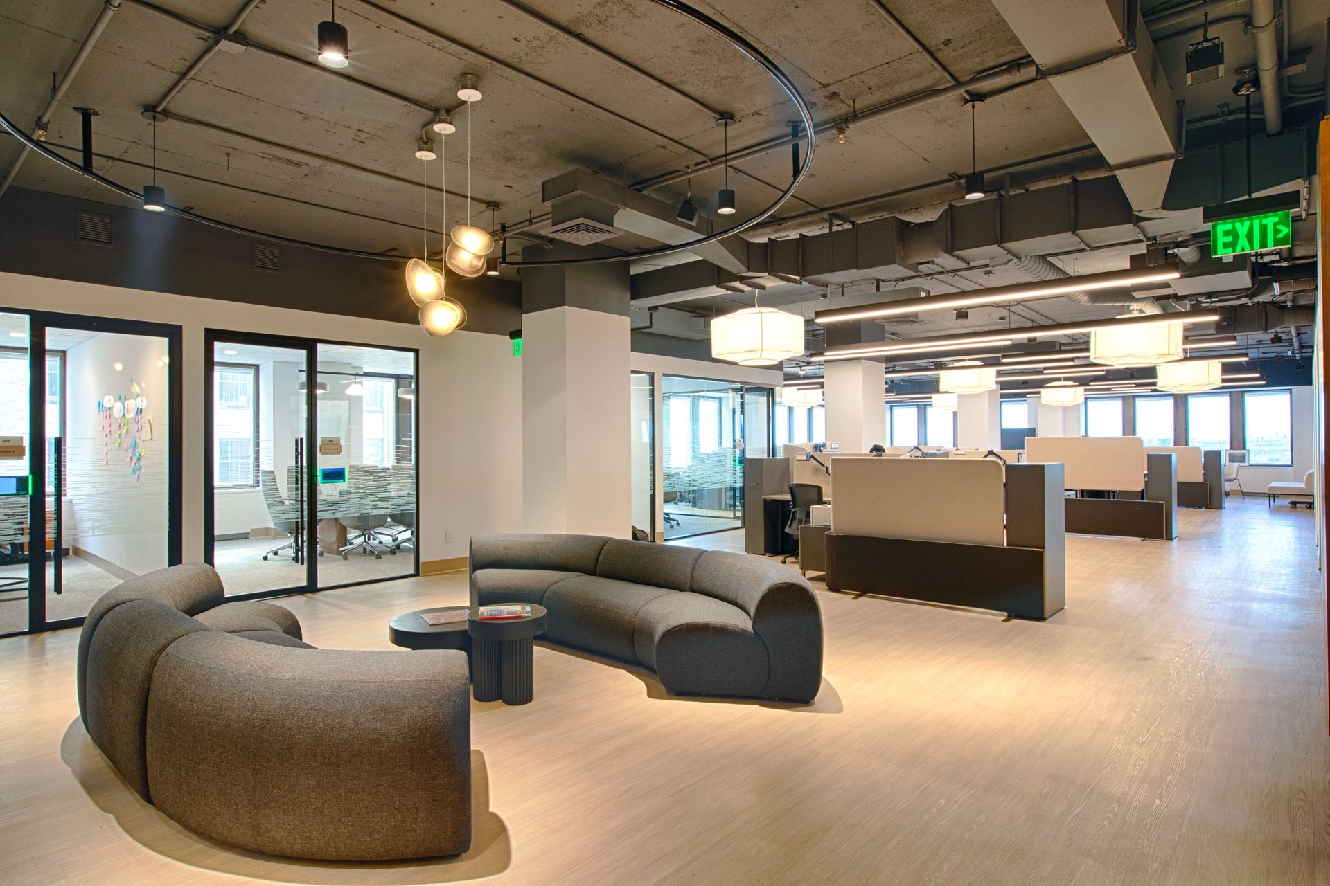 Modern office space with curved gray sofa, glass-walled offices, and rows of workstations.