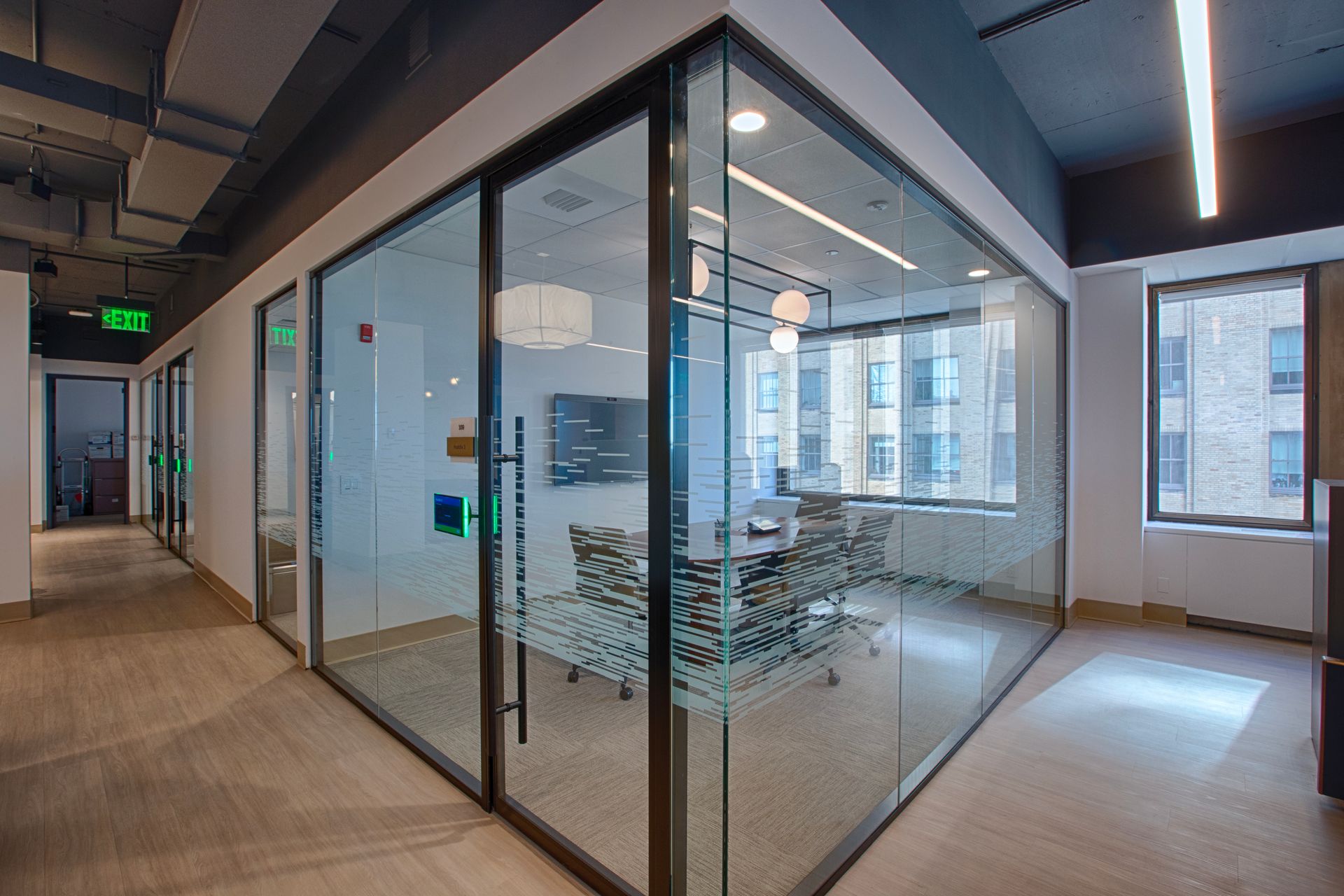 Office hallway with glass-walled conference room. A table and chairs are inside. Building windows are visible in the background.