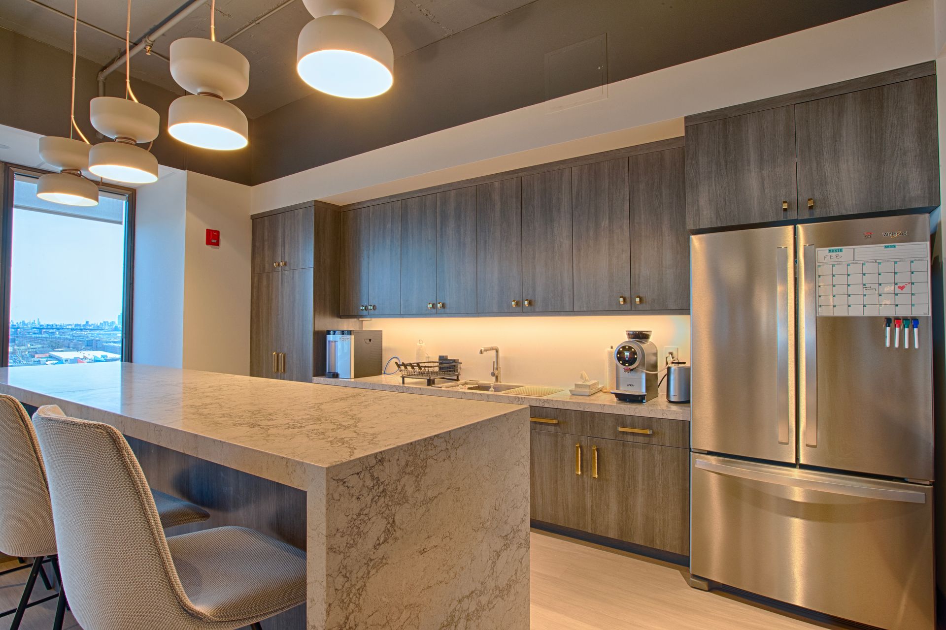 Modern office kitchen with a large island, stainless steel refrigerator, and dark cabinets.