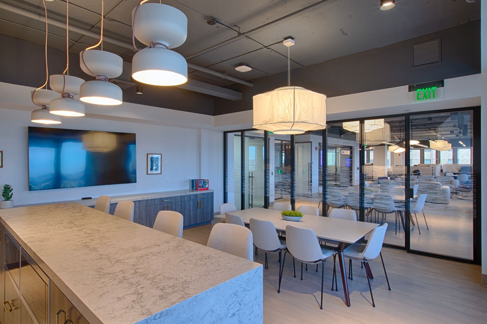 Modern office break room with long counter and dining table, glass wall to a work area.