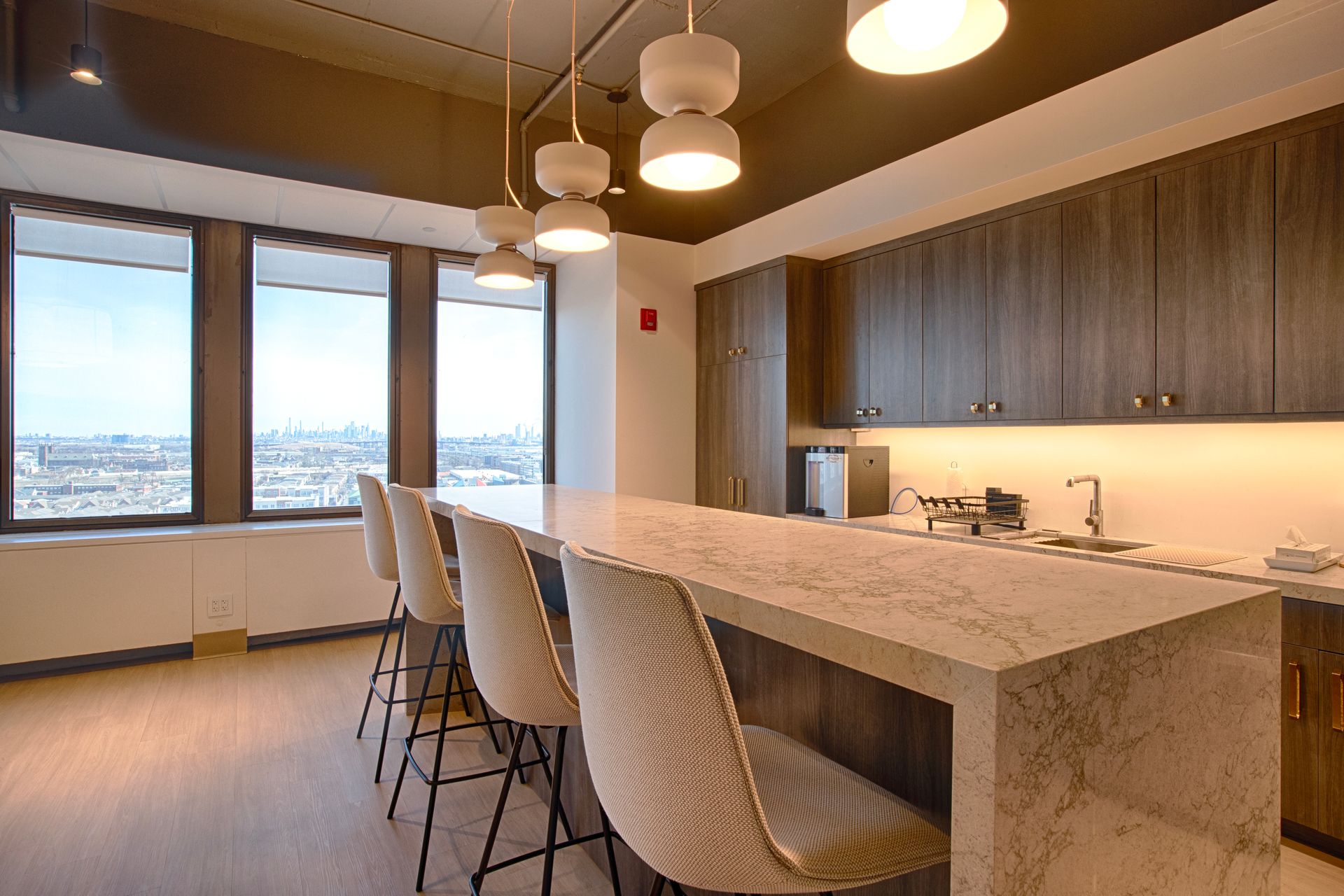 Modern office kitchen with a counter and stools, windows overlooking a city, and overhead lighting.