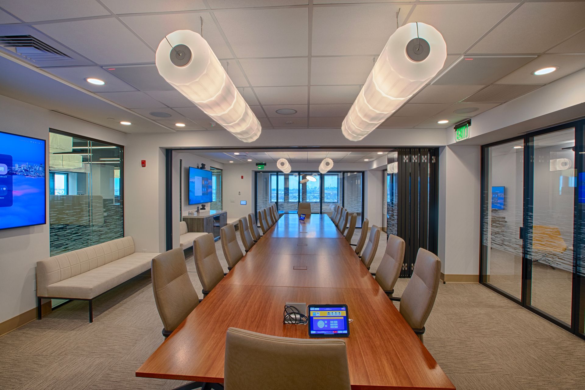 Conference room with long wooden table, beige chairs, two cylindrical lights, and glass walls.