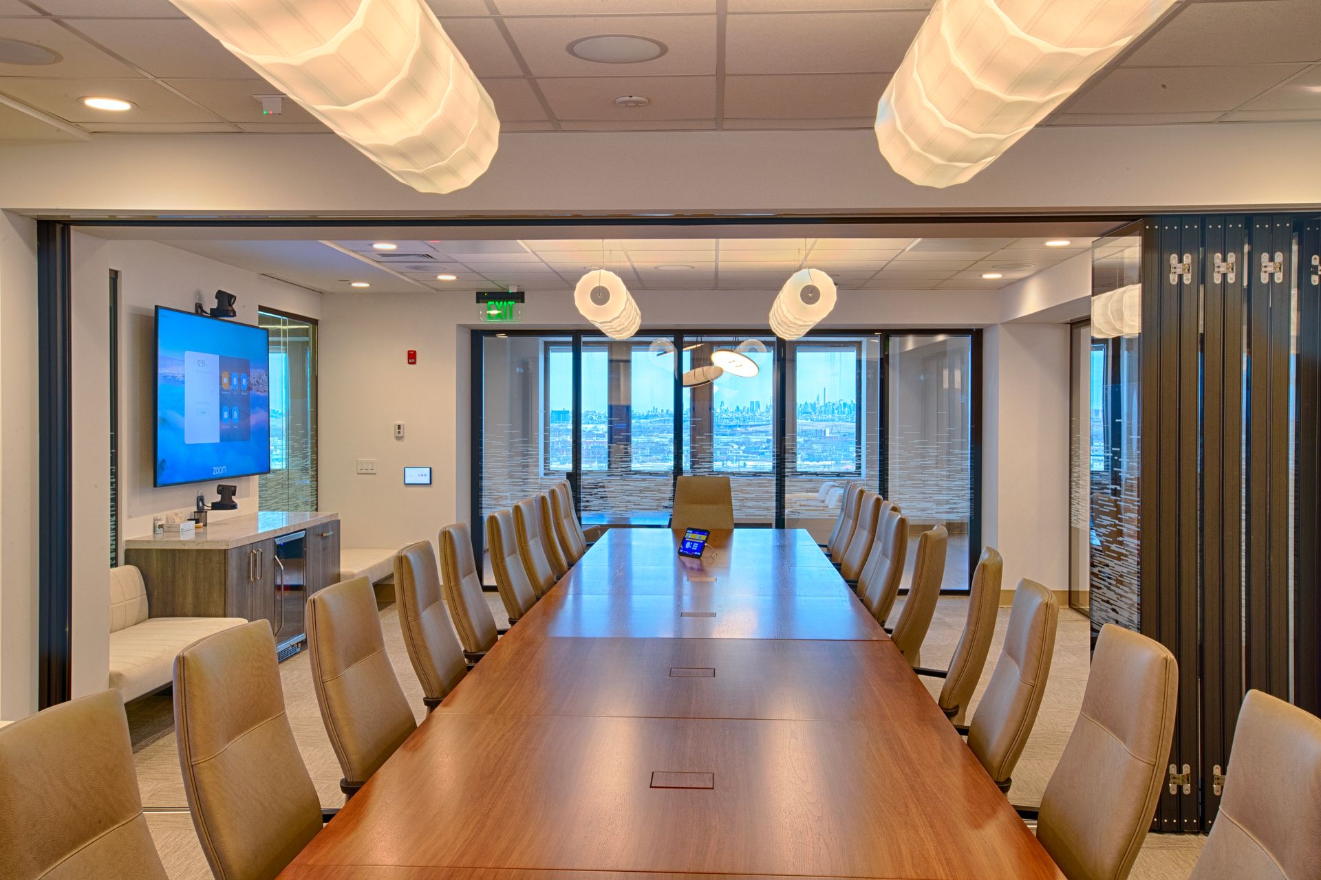 Conference room with long wooden table, tan chairs, large screen, and city view through windows.