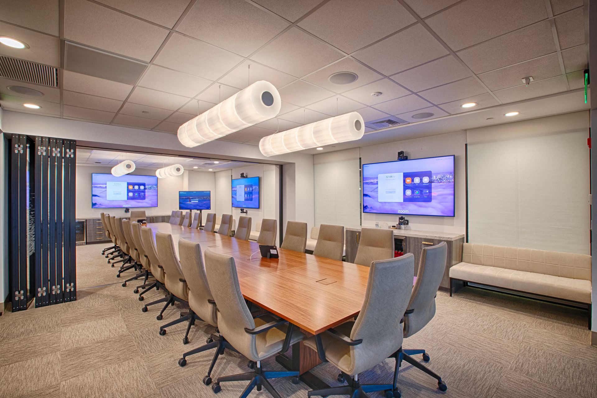 Conference room with large wooden table, chairs, screens, and modern lighting.