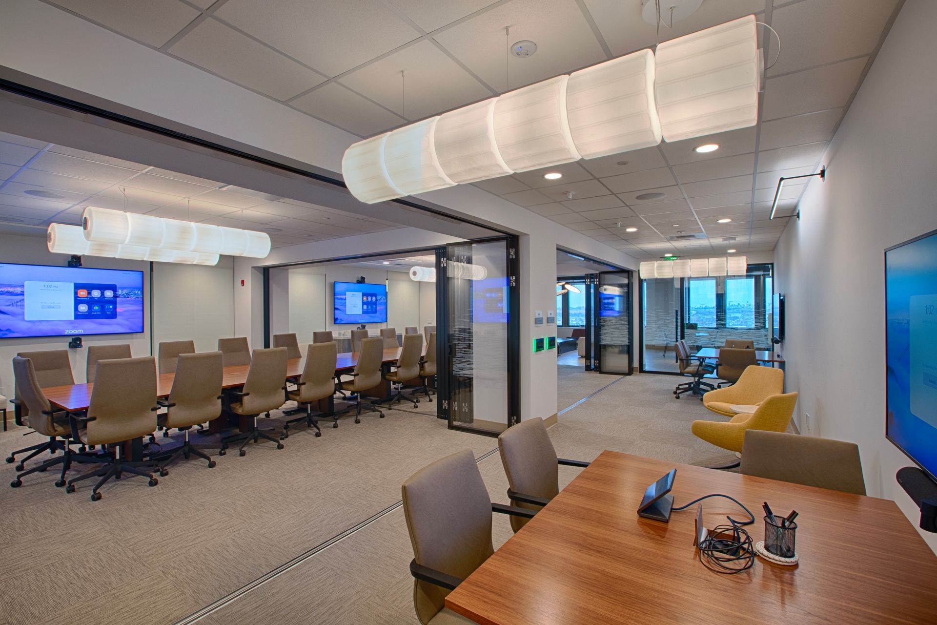 Conference room with large wooden table, chairs, and multiple screens. Beige carpet and modern lighting.