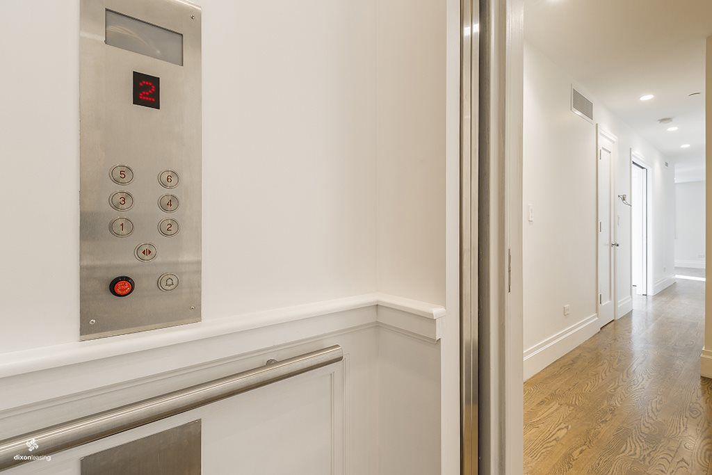 Elevator control panel on white wall, with buttons and a hallway visible.