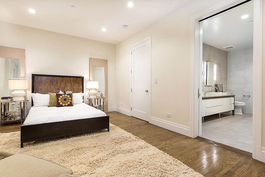 Bedroom with bed, rug, and bathroom visible through an open doorway; neutral color palette.