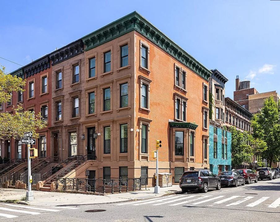 Brick apartment buildings on a city street corner, cars parked, sunny day.