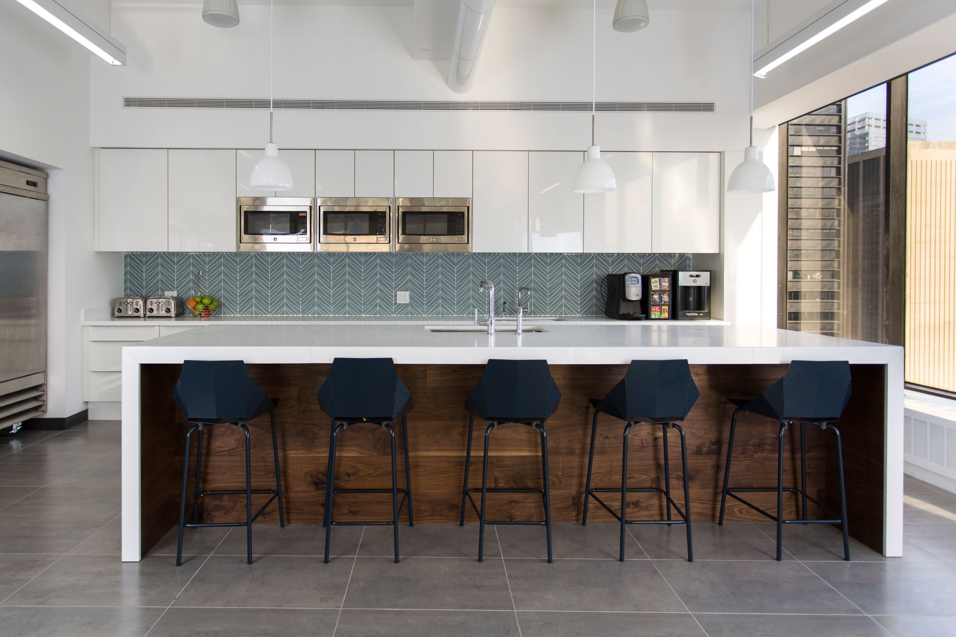 Modern office kitchen with white cabinets, blue tile backsplash, long countertop with bar stools.