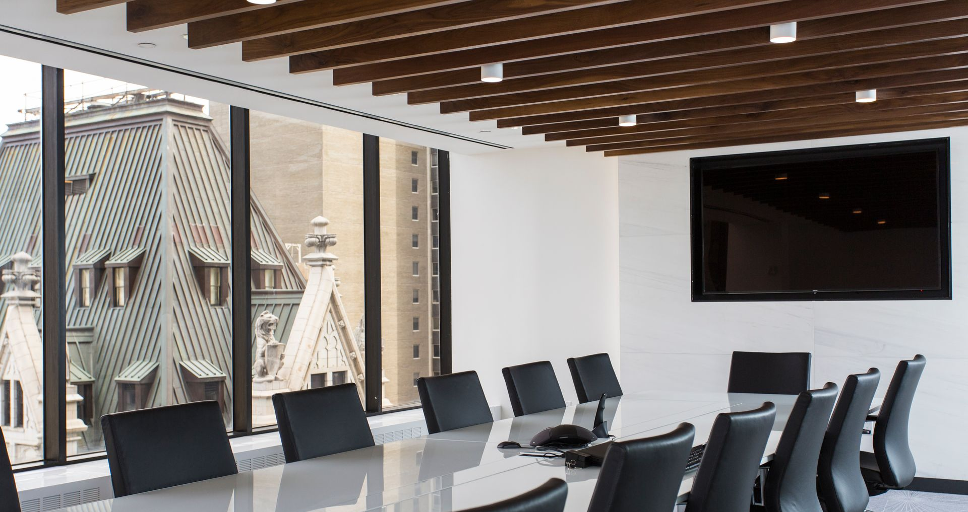 Conference room with large windows overlooking a city, long table, black chairs, and a mounted TV.