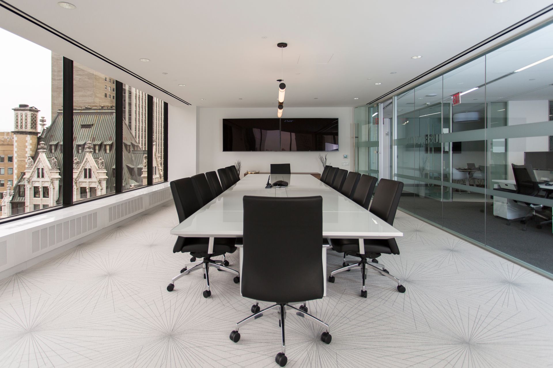 Conference room with long white table, black chairs, large windows, and an office in the background.