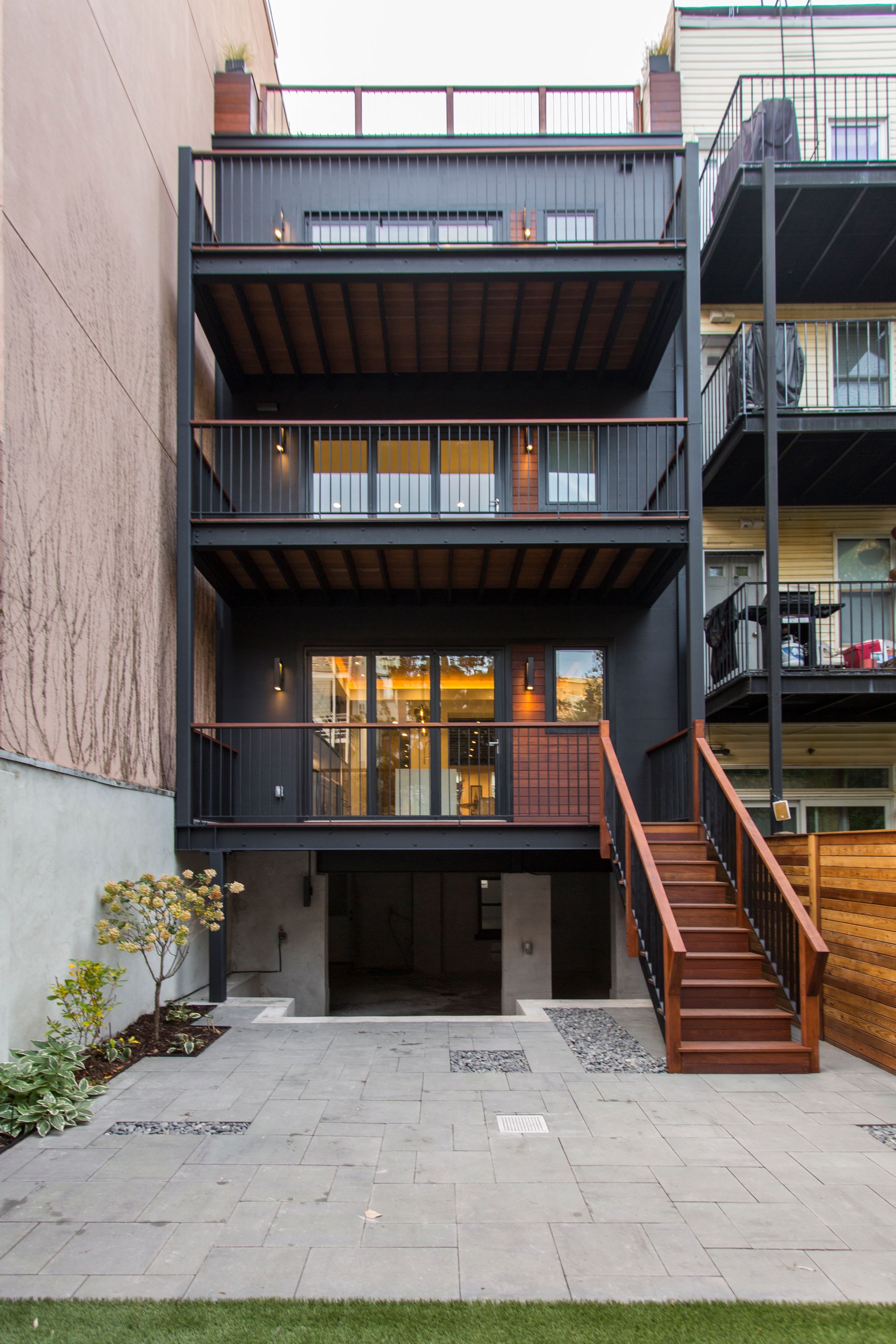 Rear exterior of a dark modern townhouse with three balconies.  Wooden stairs lead up to the upper levels and a patio below.