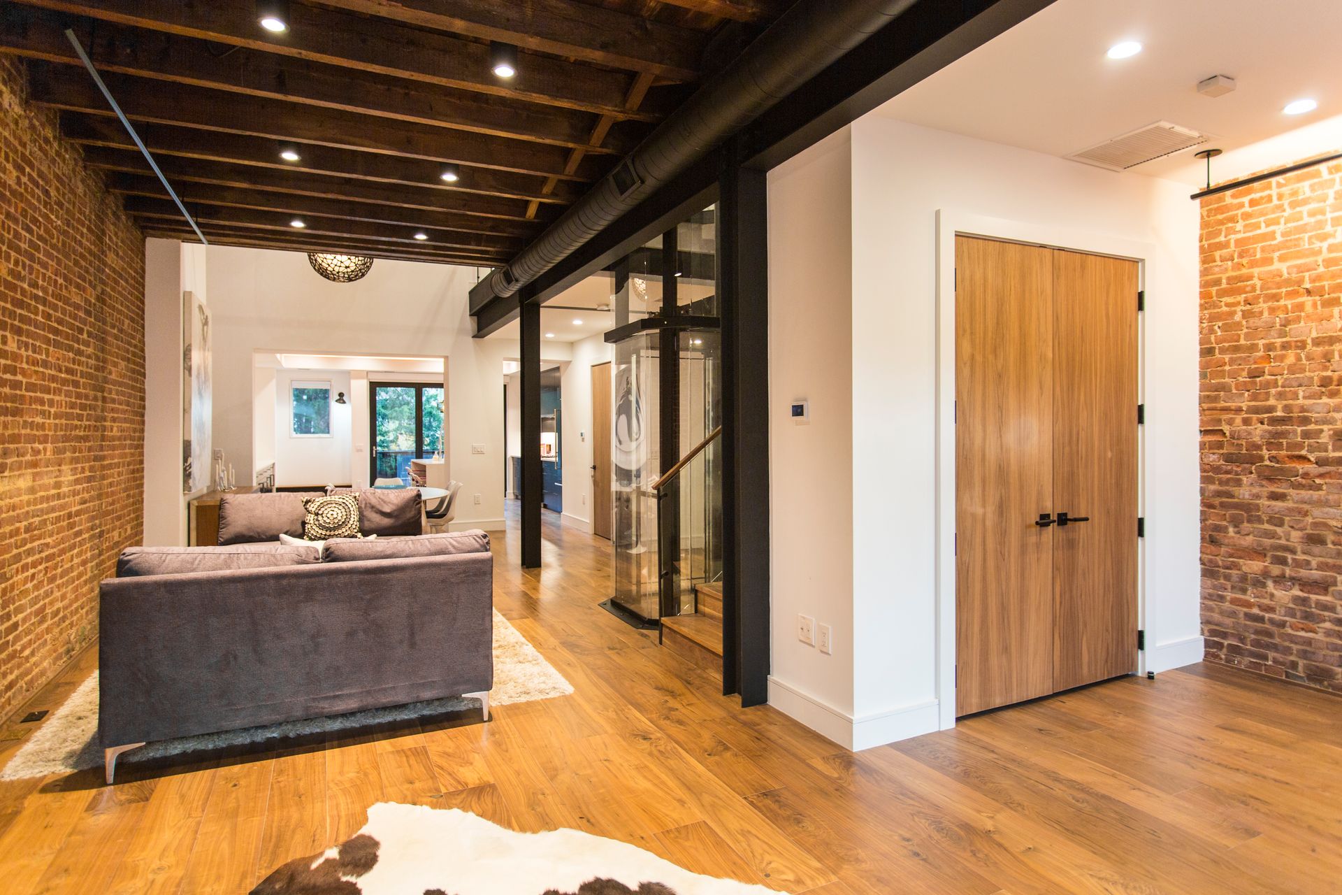 Spacious interior with exposed brick walls, wood floors, and a glass-enclosed staircase, leading to another room.
