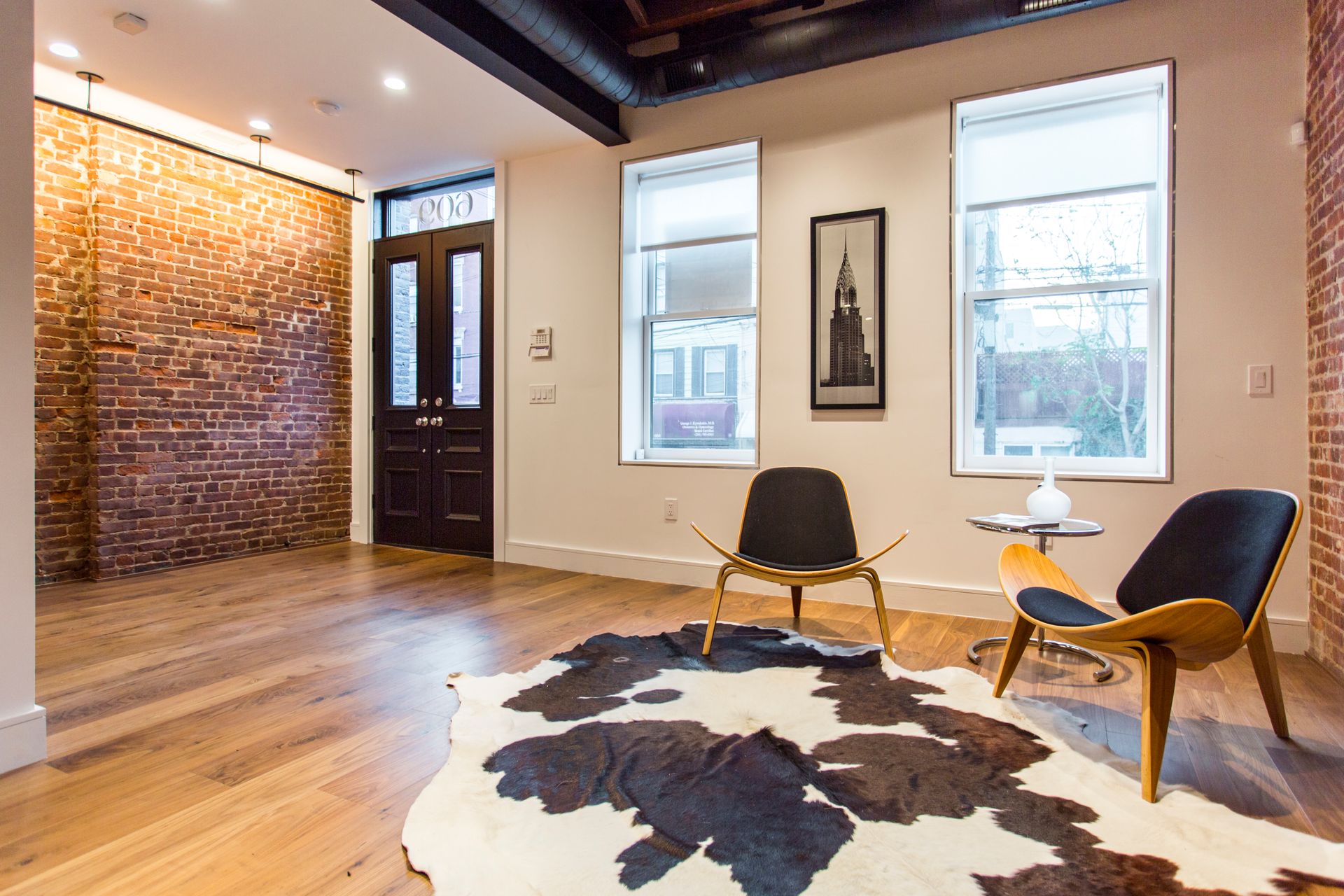 Living room with exposed brick wall, wood floor, cowhide rug, modern chairs, and three windows.