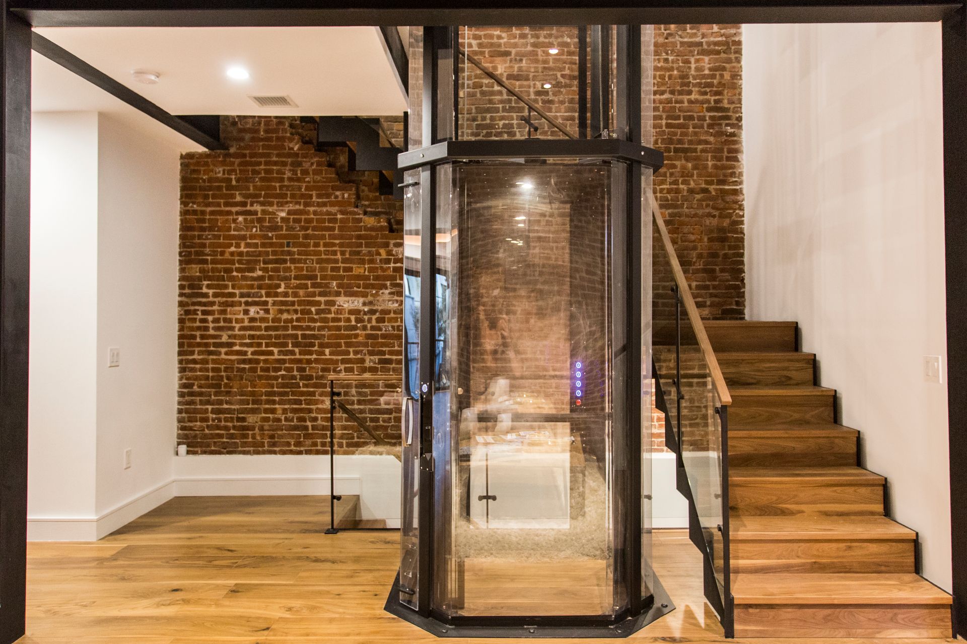 A modern interior with a glass elevator, brick wall, wooden stairs, and light wood flooring.