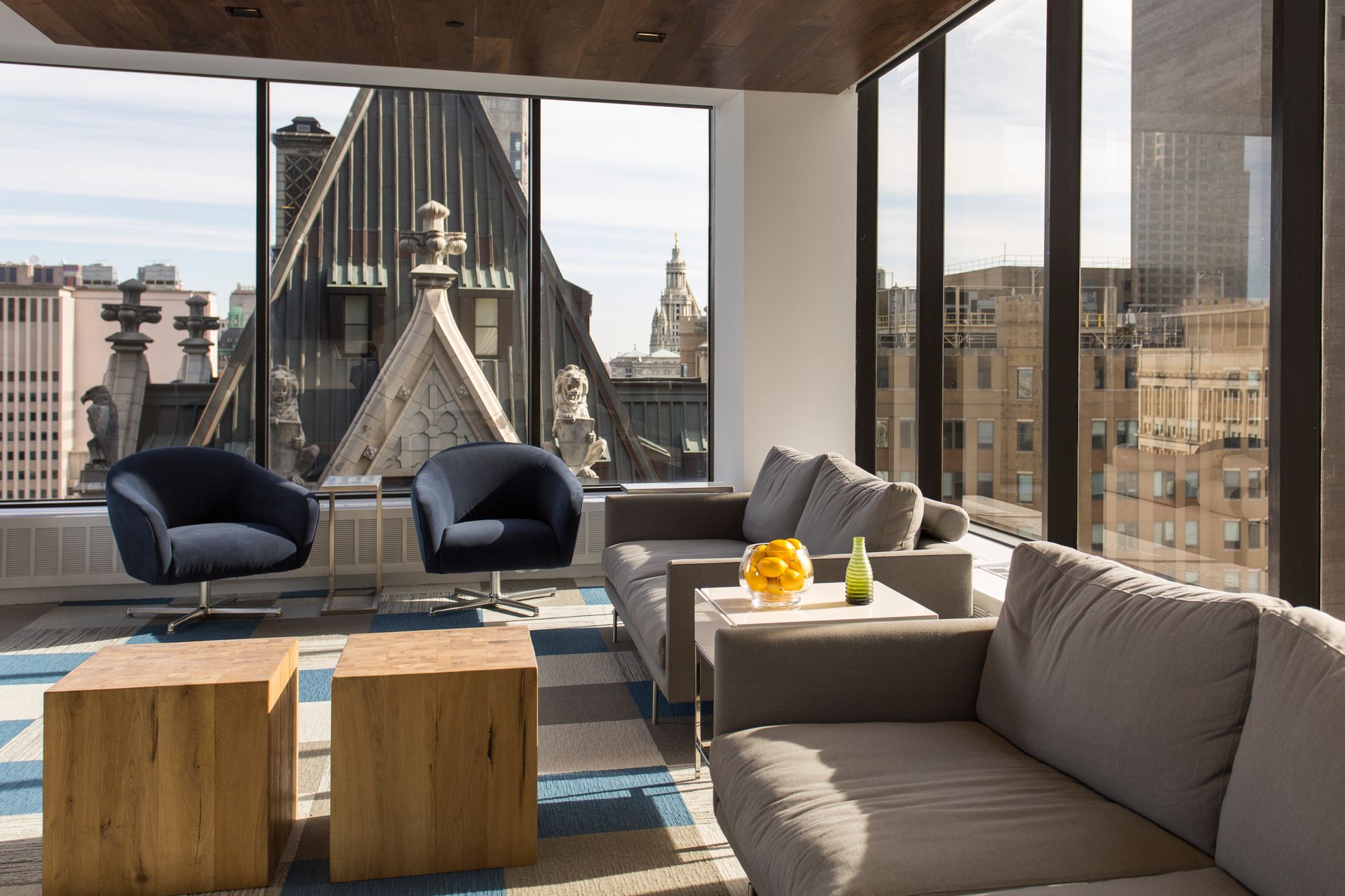 Lounge area with city view: two armchairs, two wood cube tables, gray sofas, large windows, and a cityscape backdrop.