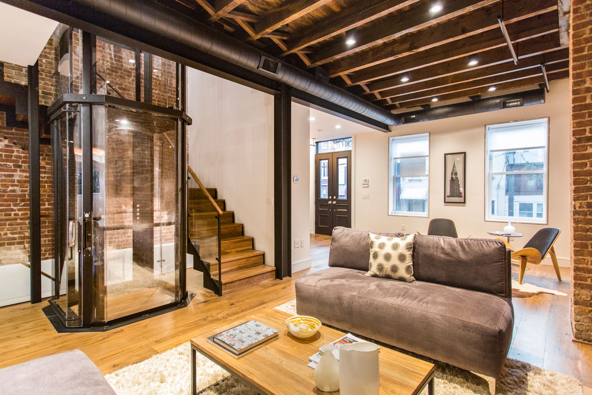 Modern living room with glass elevator and exposed brick. Sofa, stairs, and windows visible.