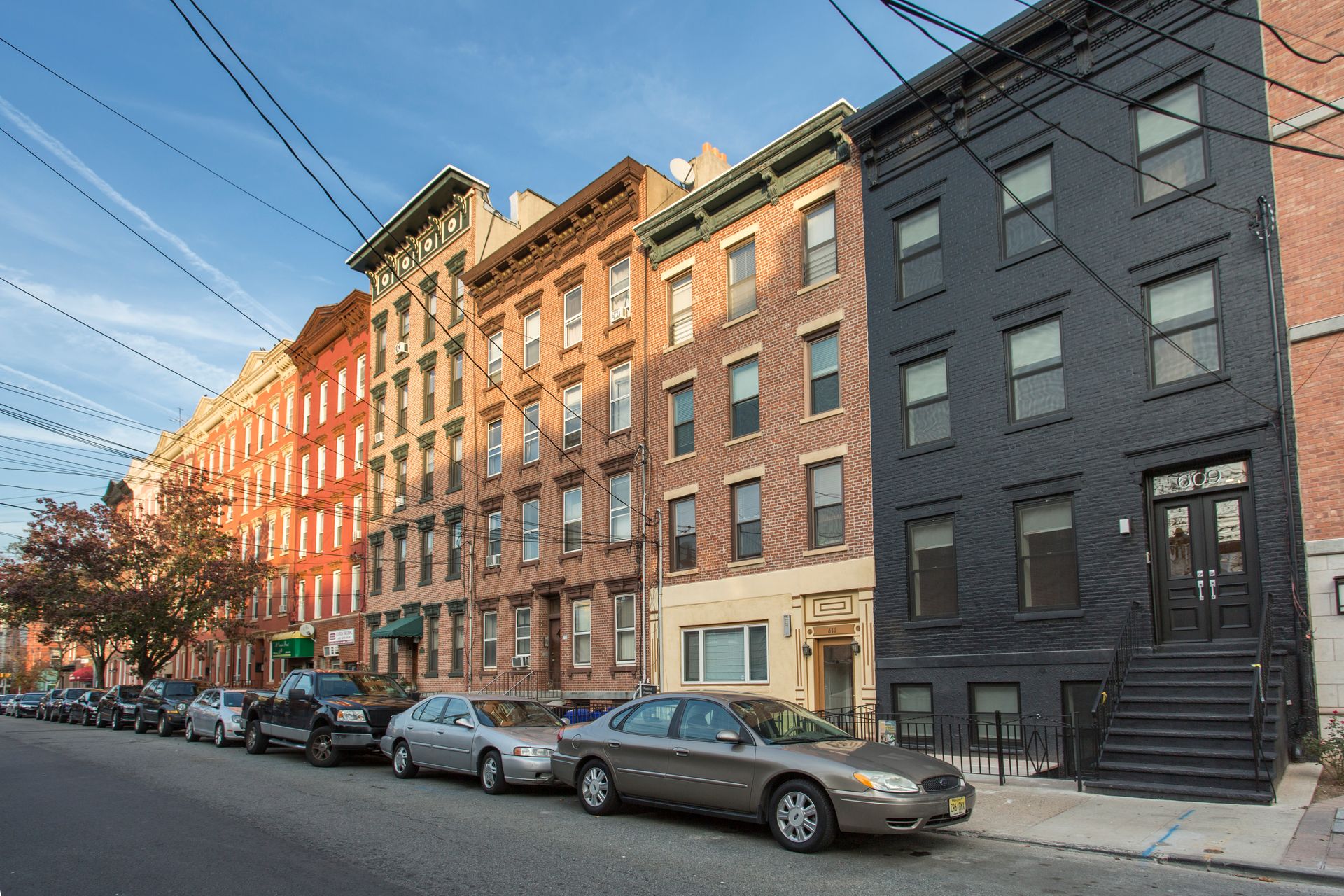 Row of brick and painted brownstone buildings on a city street, cars parked along the curb, blue sky.