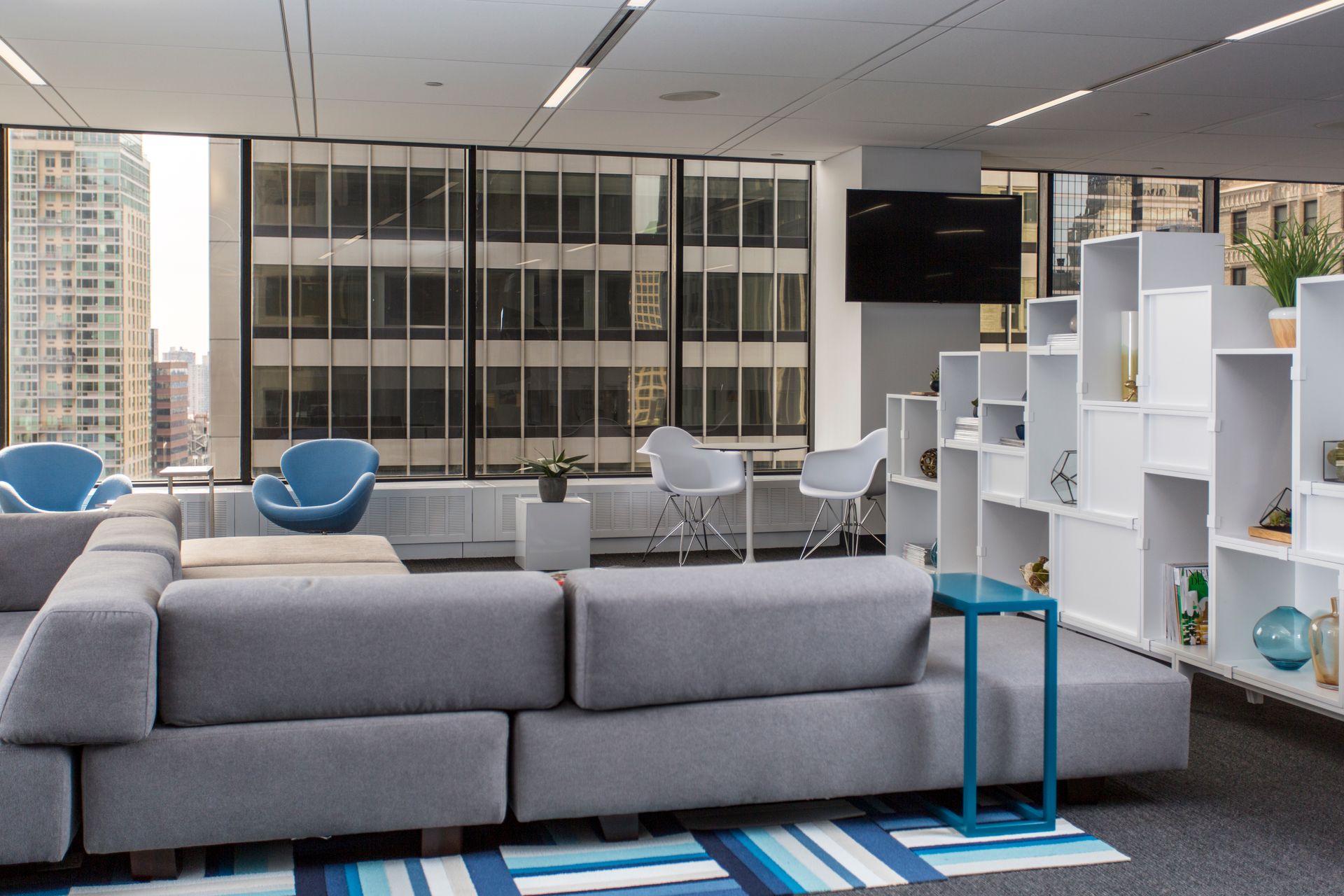 Modern office lounge area with gray sectional sofa, blue chairs, white shelving unit, and city view.