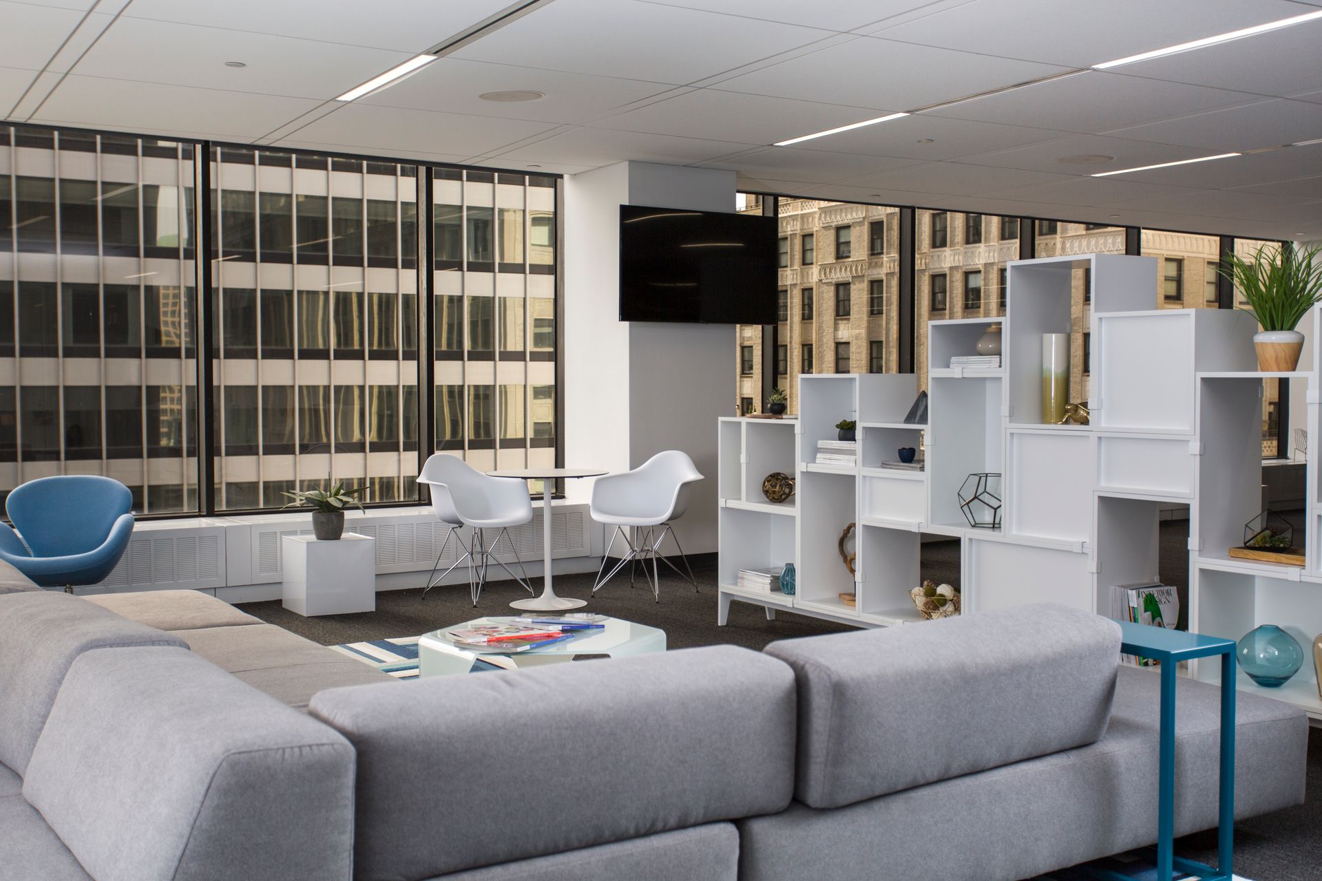 Modern office lounge with gray sectional sofa, white shelving, two white chairs, and city view.