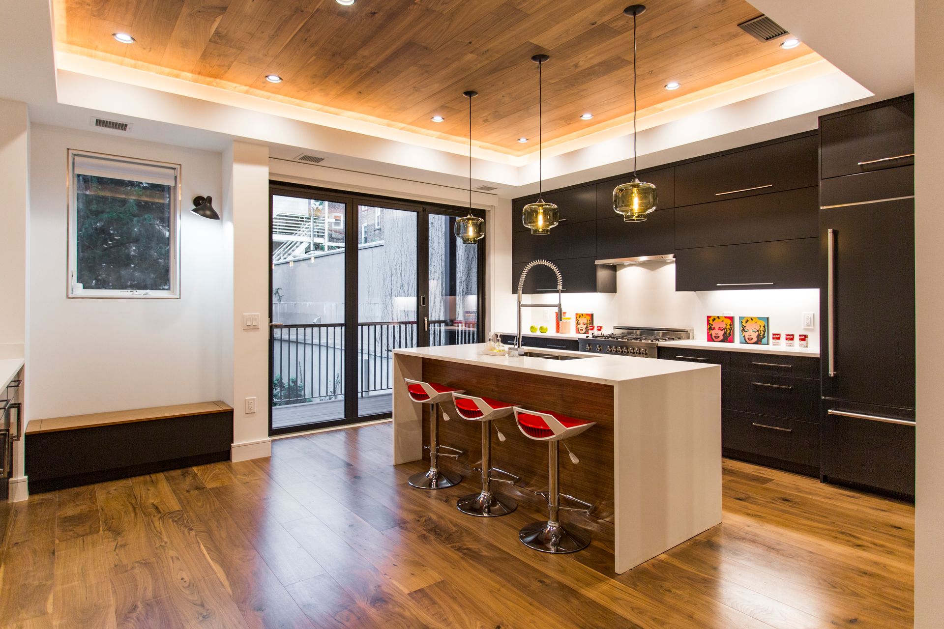 Modern kitchen with wood ceiling, island with red stools, black cabinets, and sliding doors to balcony.