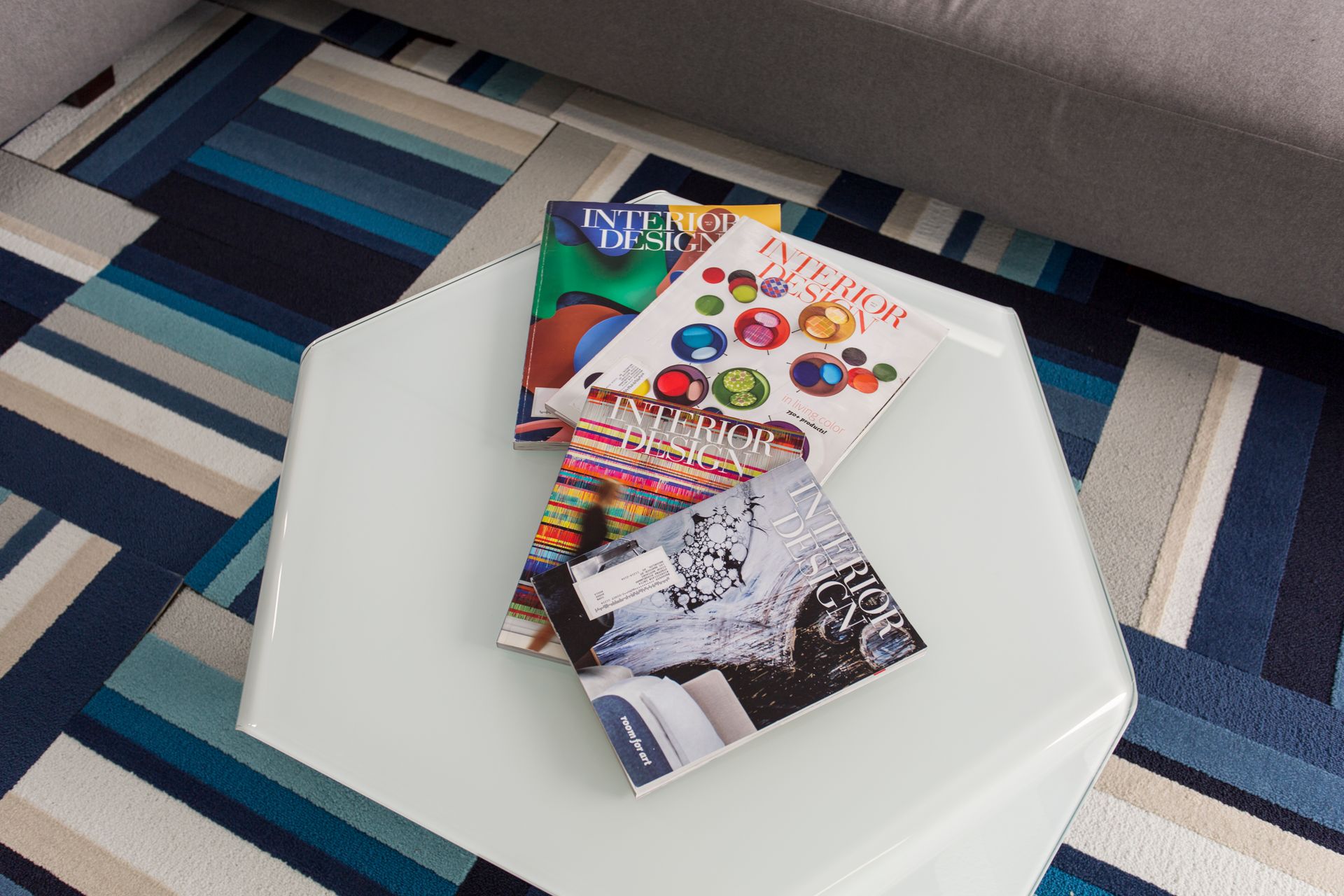 Magazines stacked on a white, geometric table. The table sits on a blue, patterned rug.