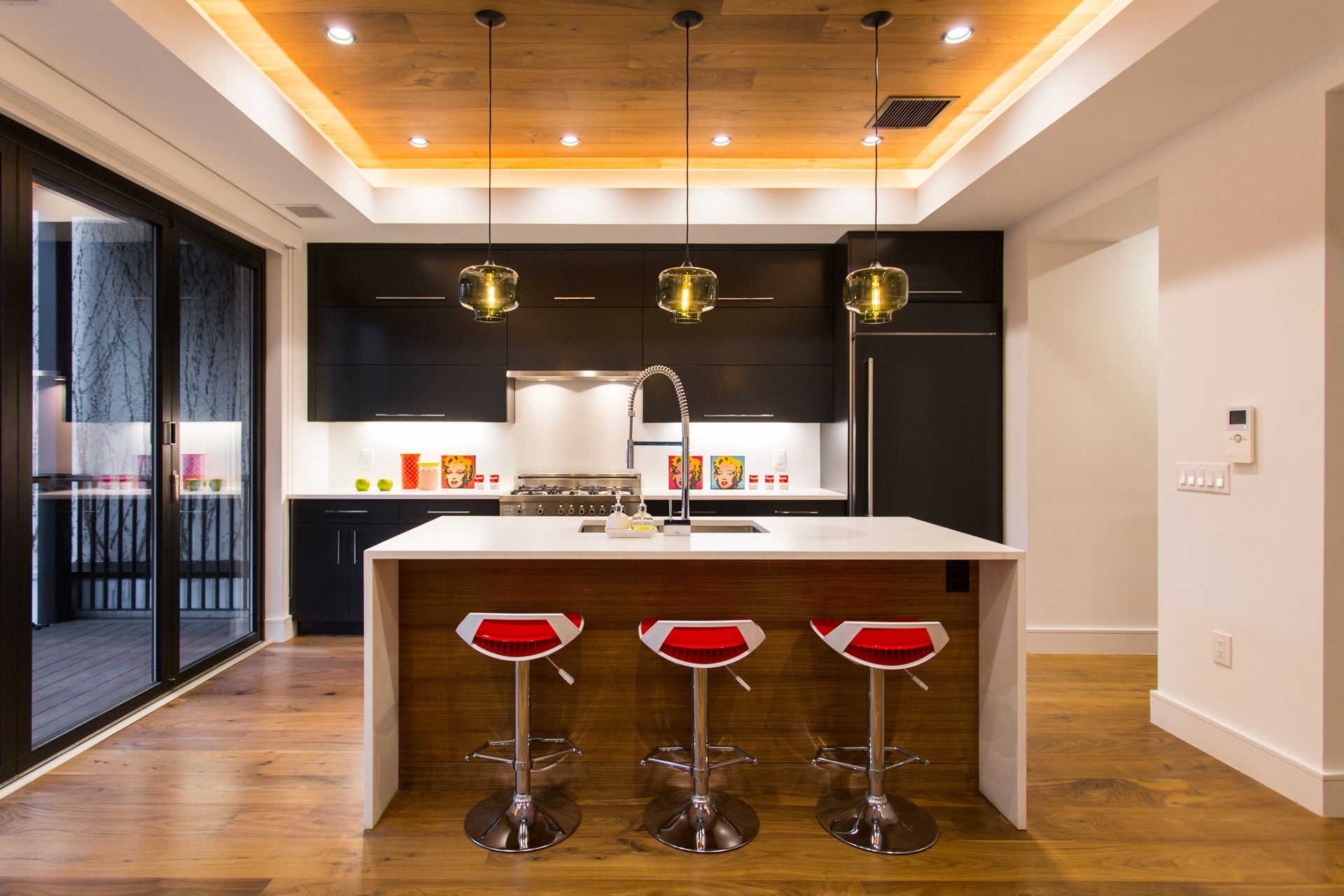 Modern kitchen with dark cabinets, white island with red stools, wood ceiling, and sliding glass doors.