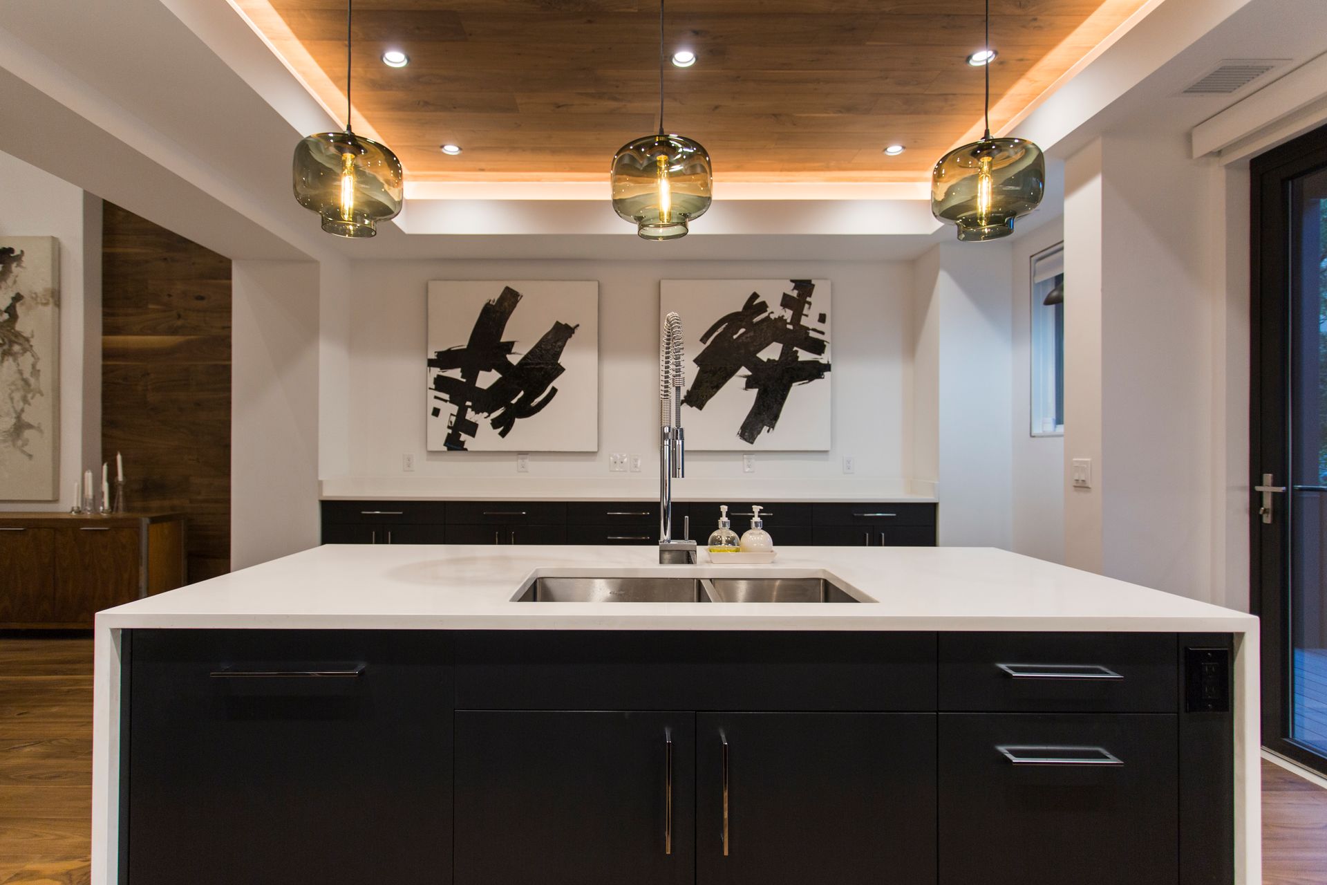 Modern kitchen with island, black cabinets, white countertop, two abstract paintings, wood ceiling, pendant lights.