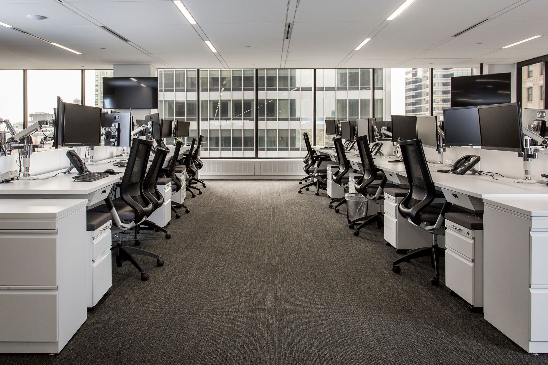 Modern office interior with rows of desks, chairs, and computer monitors. Windows overlook a city.