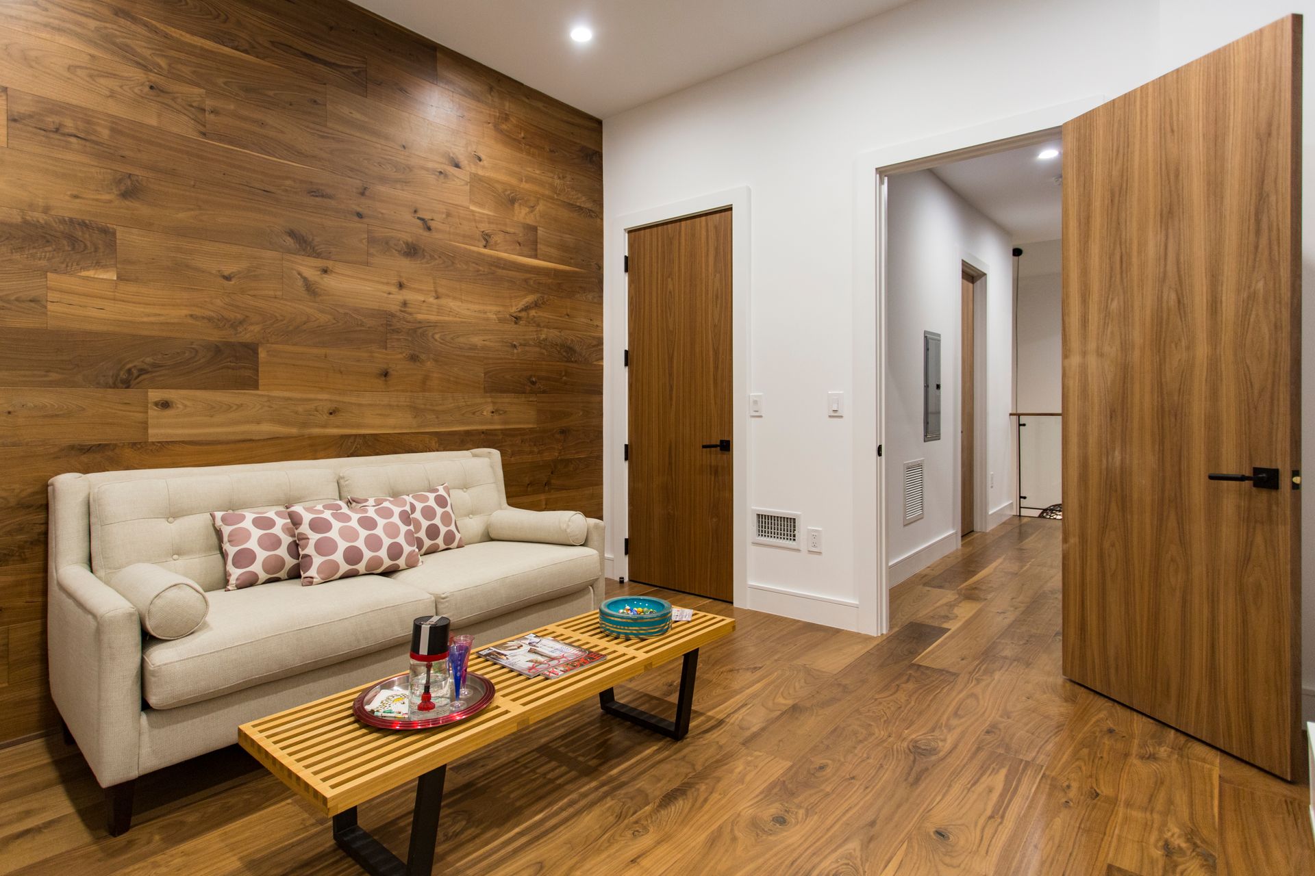 Living room with wood panel wall, beige sofa, wooden floor, open door to hallway.