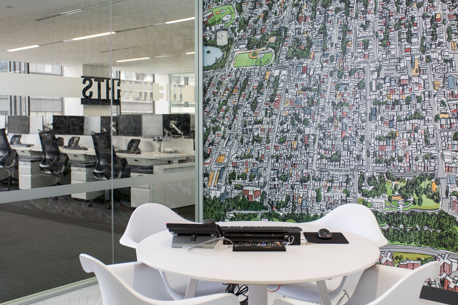 White table and chairs in front of a city map mural. Office space visible through glass, desks and computers.