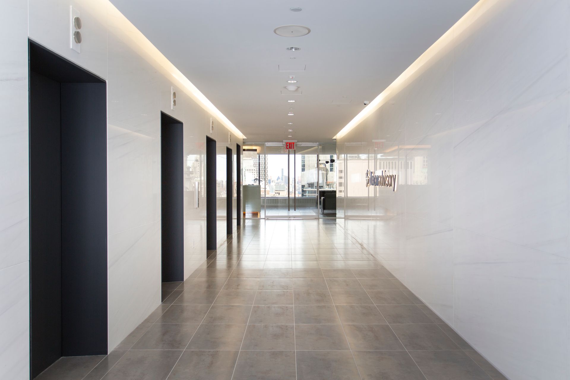 Modern hallway with elevator doors on the left, and glass doors at the end of the hall. White walls and ceiling.