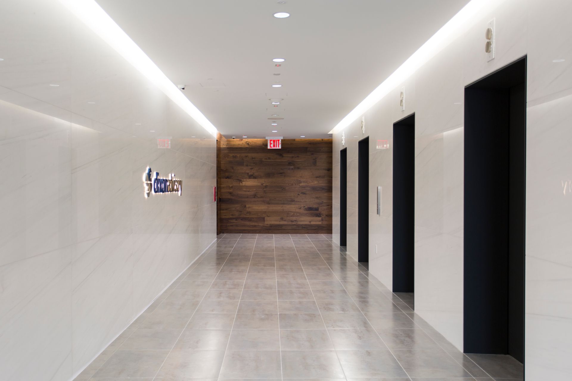 Long hallway with white walls, three dark elevator doors, wood panel wall, and exit sign.